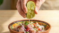 Caucasian male hand squeezing fresh lime juice over ceviche dish with raw fish, red onion and cilantro in wooden bowl for seafood preparation and cooking.