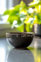 Black ceramic bowl on reflective surface with soft green bokeh background from window plant creates serene minimalist composition for wellness and spa concepts.