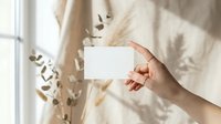 Female hand holding blank white business card mockup with eucalyptus shadow overlay on beige fabric background for branding presentation.