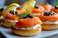 Mini bagels topped with cream cheese, smoked salmon, black caviar, fresh dill and lemon slices arranged on white plate, close up view of gourmet appetizers.