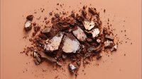 Crushed bronzer powder with shimmering particles scattered on warm beige background, creating texture for cosmetic product photography.