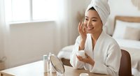 Young Asian woman in white bathrobe and towel applying skincare cream while looking in mirror, morning beauty routine in bright bedroom spa atmosphere.
