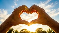 Hands forming heart shape against sunset sky with warm golden light shining through fingers creating romantic silhouette for love and affection concepts.