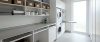 Modern white laundry room with stacked washer dryer, built-in storage shelves, folding counter and natural light from large window.