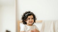 Young child with curly dark hair and medium skin tone wearing white shirt, smiling at camera in bright indoor setting. Suitable for family, parenting, childcare, and lifestyle marketing materials.