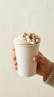Hand holding white paper cup with hot chocolate topped with whipped cream and mini marshmallows against neutral beige background for cozy winter beverage concept.