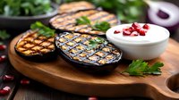 Grilled eggplant slices with charred grill marks served on wooden board alongside fresh yogurt dip garnished with pomegranate seeds and parsley leaves.
