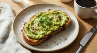 Creamy avocado toast topped with fresh herbs and seeds on artisan bread, served on ceramic plate with coffee for healthy breakfast or brunch meal.