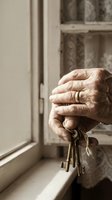 Elderly hands holding vintage keys by window with wedding ring, symbolizing home ownership, memories, inheritance, and life transitions in warm natural light.