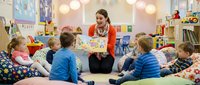 Female educator with brown hair wearing red cardigan leading interactive storytelling session with diverse group of young children seated in circle during classroom activity in bright, colorful learning environment.
