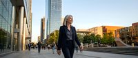 Blonde woman in dark business suit walking through modern urban plaza with skyscrapers and contemporary architecture during daytime, professional corporate environment.