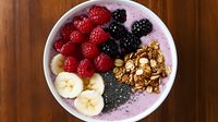 Fresh acai bowl topped with sliced banana, raspberries, blackberries, granola and chia seeds on wooden background, healthy breakfast smoothie bowl.