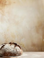 Rustic artisan sourdough bread loaf with flour dusting on light concrete surface against neutral beige wall background, minimalist food photography style.