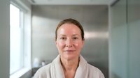 Middle-aged Caucasian woman in white bathrobe smiling in modern bathroom interior with natural lighting for wellness and self-care concepts.