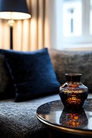 Elegant glass vase with amber reflections on modern coffee table, navy blue velvet cushions and warm lamp lighting create cozy living room atmosphere.