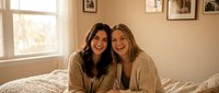 Two young women friends laughing together on bed in cozy bedroom with warm natural light and framed photos on wall creating intimate friendship moment.