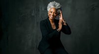 Senior woman with short white hair, wearing black blazer, smiling while talking on smartphone against dark textured background. Professional communication portrait.