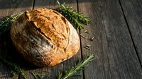 Freshly baked artisan sourdough bread loaf with golden crust and rosemary herbs on rustic wooden table for bakery and cooking concepts.