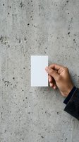 Hand holding blank white card against textured concrete wall surface, ideal for business card mockups, branding presentations, and design templates.