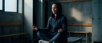 Young African American woman in dark clothing sitting cross-legged in meditation pose in modern industrial loft space with natural window lighting.