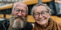 Happy elderly Caucasian couple with glasses smiling warmly outdoors, mature man with full gray beard and woman in cozy scarf enjoying retirement together.