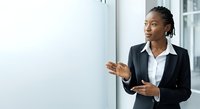 Young African American female professional in business attire presenting ideas at whiteboard during corporate meeting or training session in modern office environment.