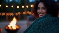 Young African American woman wrapped in cozy knit blanket smiling warmly by outdoor fire pit with string lights creating magical evening atmosphere.