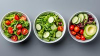 Fresh vegetable salads in white bowls with tomatoes, cucumber, avocado and mixed greens on gray background, healthy lunch concept, top view.