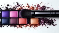 Makeup palette with vibrant eyeshadow colors in purple, pink, and coral shades, professional black brush, scattered powder texture on white background.