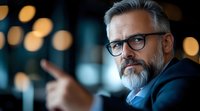 Mature Caucasian businessman with gray beard and glasses in modern office setting with bokeh lights background for corporate presentations.