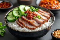 Sliced roasted duck breast with chili sauce served on white rice in black ceramic bowl, garnished with fresh cucumber and green onions. Asian cuisine.