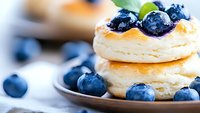 Fresh blueberries atop fluffy stacked pancakes with golden syrup drizzle on ceramic plate, extreme close up showing texture and layers in morning light.