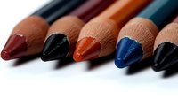 Macro shot of colorful cosmetic lip and eye pencils arranged in row showing sharpened tips against white background, suitable for beauty and makeup advertising.