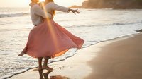 Young woman in flowing pink dress dancing freely on sandy beach at golden hour sunset with ocean waves and warm sunlight creating romantic coastal atmosphere.