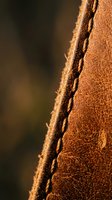 Close-up macro shot of brown leather edge with detailed stitching and natural grain texture. Warm lighting highlights craftsmanship and material quality.