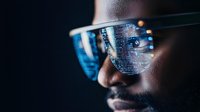 Young African American man wearing smart glasses with digital code reflection, representing augmented reality and future technology in dark setting.