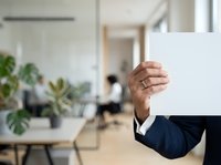 Middle-aged man with medium brown skin tone wearing a dark navy blazer holds a blank white sign in a modern office environment with colleagues and plants visible in background.
