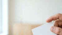 A hand holding a blank white card or paper against a soft, blurred neutral background, suitable for business, greeting, or promotional messaging.