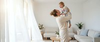 Young Caucasian mother lifting laughing toddler high in bright modern living room, sharing joyful moment together in cozy home interior.