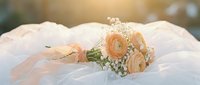 Elegant bridal bouquet with peach ranunculus and baby's breath resting on white tulle fabric in soft golden sunlight.