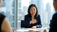 Asian female executive in dark blazer conducting business meeting in modern high-rise office with city skyline view, discussing strategy with colleagues.