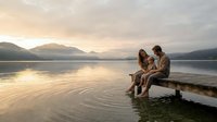 Young couple sitting on wooden dock at sunset lake with mountains, romantic moment together enjoying peaceful evening by calm water.