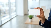 Man in business attire holding blank white card in modern office with large windows, suitable for corporate branding, business communication, and professional marketing materials.