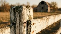 Weathered wooden fence post with rustic barn in blurred background during golden hour sunset light creating warm agricultural countryside atmosphere.