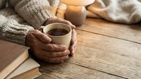 Cozy autumn morning scene with hands holding warm coffee mug, knitted sweater, books and candles on rustic wooden table creating hygge atmosphere for relaxation.