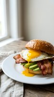 Gourmet breakfast burger with crispy bacon, fried egg, avocado slices on sesame seed bun served on white plate with natural window lighting for food photography.