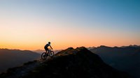 Mountain biker riding on rocky summit at sunset with dramatic alpine landscape and golden sky in background for adventure and outdoor sports content.