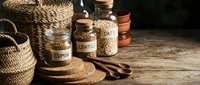 Labeled glass jars containing dried legumes and grains like lentils and oats arranged on wooden surface with woven baskets and measuring spoons for cooking and meal preparation.