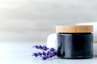 Black glass cosmetic jar with wooden lid and fresh lavender sprig against light background, minimalist spa and beauty product photography styling.