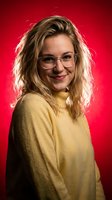 Young blonde Caucasian woman wearing glasses and yellow sweater smiling confidently against vibrant red background for professional headshot photography.